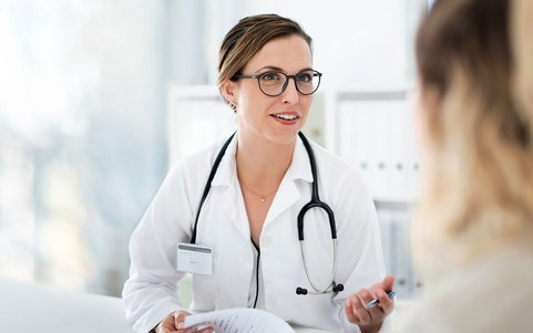 Eine Ärztin im weißen Kittel mit Stethoskop sitzt einem Patienten gegenüber und hört während eines Beratungsgesprächs zu